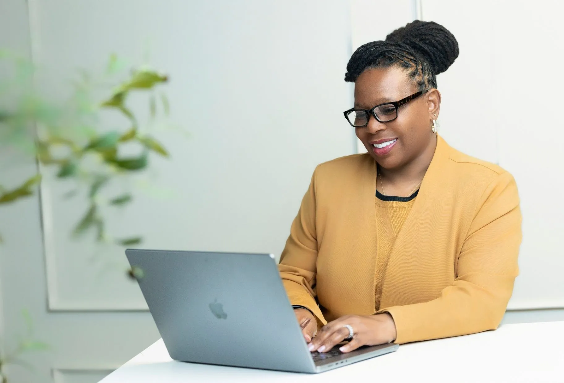 Dr. Darlene Williams-Prades smiling while working remotely at a white desk with a laptop, wearing glasses and a mustard blazer